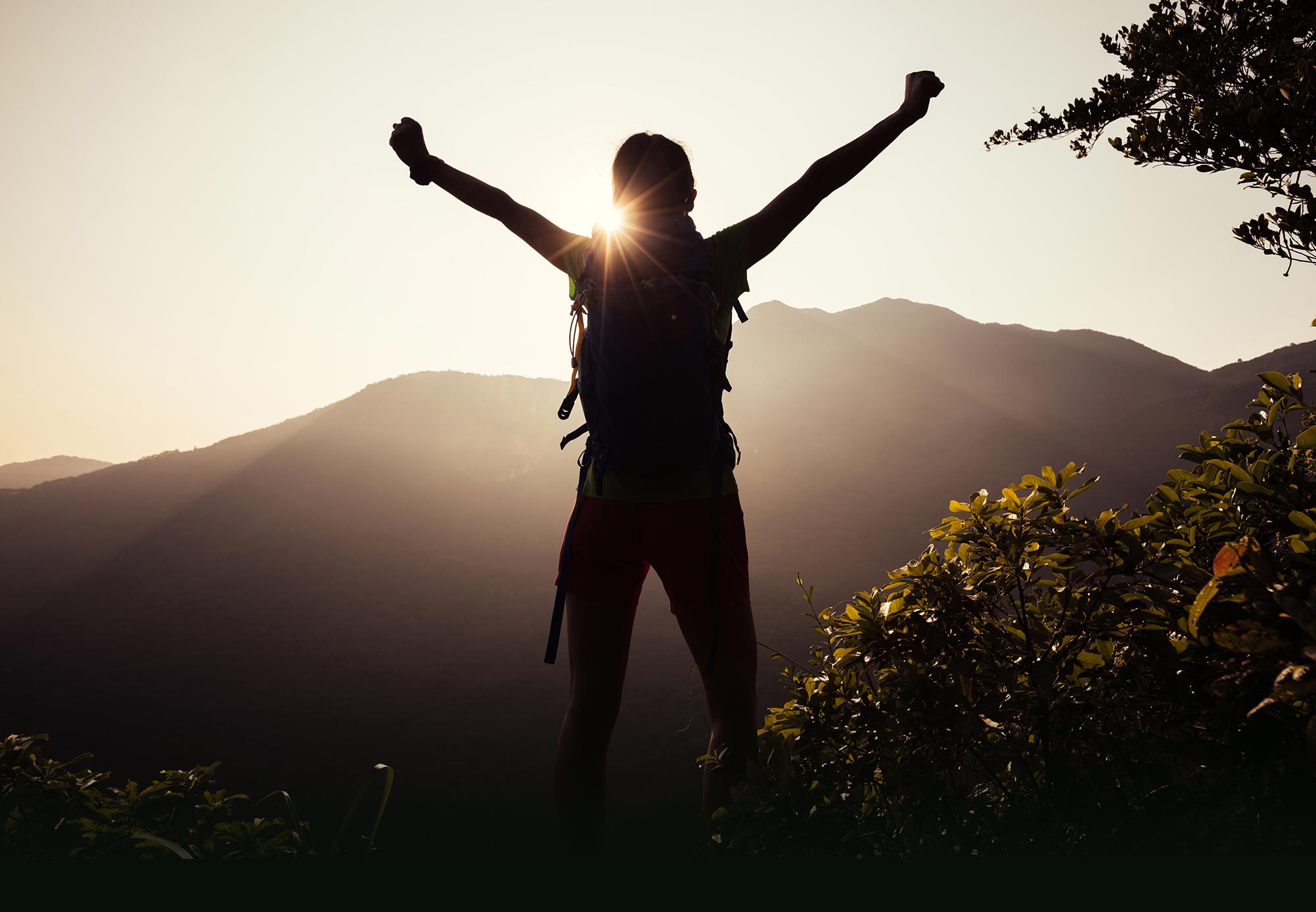 Person with arms raised silhouetted against setting sun and mountain peak